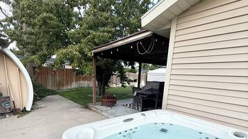 Un jacuzzi en un patio trasero con un porche cubierto y una valla de madera al fondo.