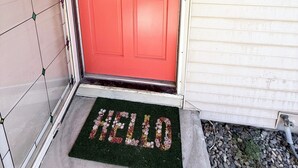 Uma porta vermelha com a placa 'WELCOME SUCCAS', um tapete de entrada com a palavra 'HELLO' em letras florais, e uma caixa de correio à direita.