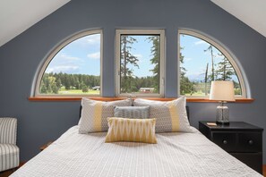 Ein Schlafzimmer mit einem großen Bett, zwei Fenstern, einem Nachttisch und einem Sessel.