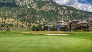 Un terrain de golf avec un mât de drapeau au premier plan, entouré d’une pelouse bien entretenue. Au fond, on aperçoit des immeubles à plusieurs étages dotés de balcons, ainsi qu’un paysage montagneux.