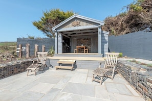 A covered outdoor seating area with wooden chairs and a table, surrounded by stone tiles and a stone wall.