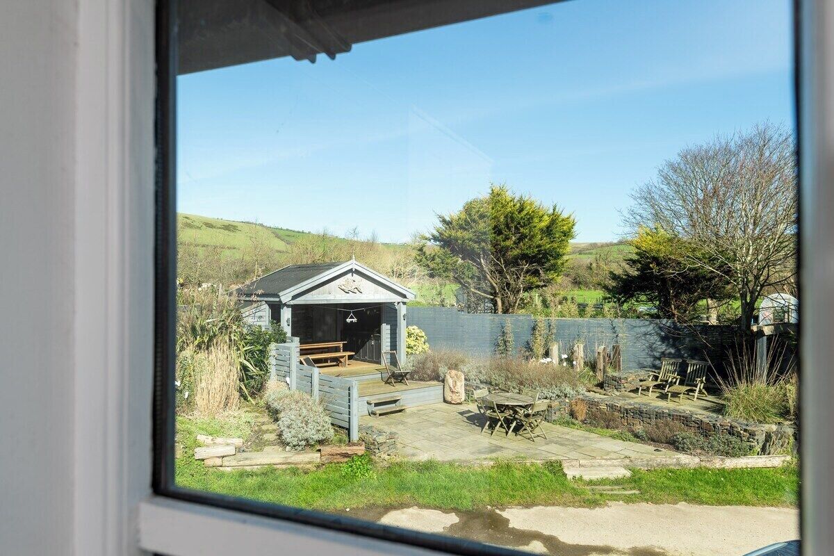 A view through a window of a house with a patio area, a small shed, and a fenced garden.