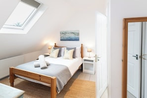 Una camera da letto con finestra a lucernario, un letto con cuscini, un quadro al muro e un comodino con una lampada.