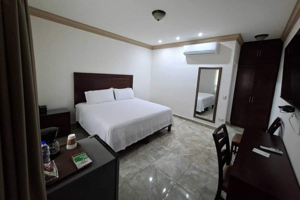 A hotel room with a bed, a desk with a chair, a mirror, a wardrobe, and an air conditioning unit.