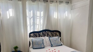 Ein Schlafzimmer mit einem Bett, Vorhängen, einem kleinen Tisch mit einer Pflanze und einem Fliesenboden.