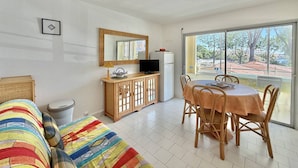 Ein Zimmer mit einer Couch, einem Esstisch mit Stühlen, einem Fernseher auf einem Schrank und einem großen Fenster mit Blick auf Bäume und Gebäude.