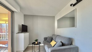 Ein modernes Wohnzimmer mit einem grauen Sofa, einem kleinen schwarzen Tisch mit einer Pflanze, einem an der Wand montierten Fernseher und einem großen Fenster.