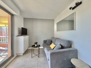 Ein modernes Wohnzimmer mit einem grauen Sofa, einem kleinen schwarzen Tisch mit einer Pflanze, einem an der Wand montierten Fernseher und einem großen Fenster.