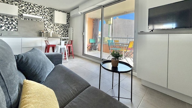 Ein modernes Wohnzimmer mit einem grauen Sofa, einem kleinen runden Tisch mit Pflanze, einer Küchenzeile mit weißer Arbeitsplatte und einem Balkon mit bunten Stühlen.