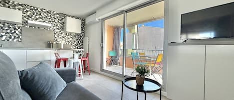 Una sala moderna con un sofá gris, una pequeña mesa redonda con una planta, una zona de cocina con encimera blanca y un balcón con sillas de colores.