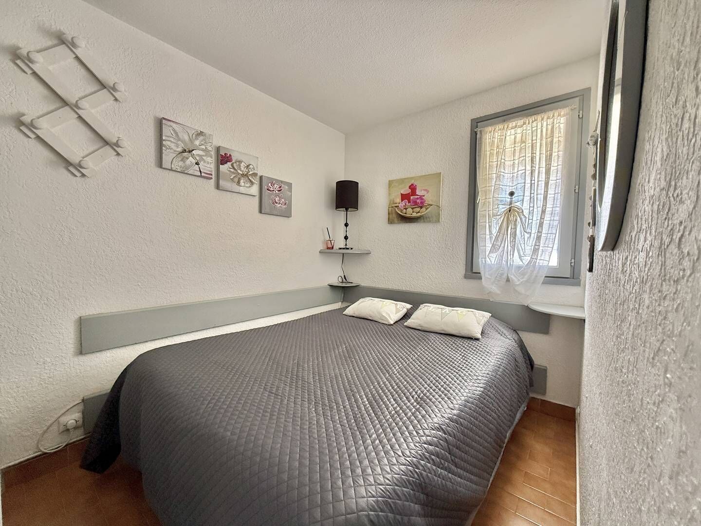 Ett sovrum med en säng, ett fönster med gardiner, ett nattduksbord med en lampa och konst på väggen.