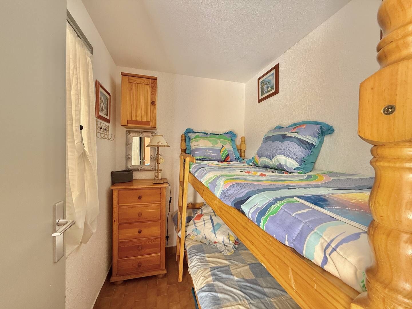 Une petite chambre à coucher accueillante, avec un lit simple, une commode en bois, un miroir et une porte munie d’un rideau.