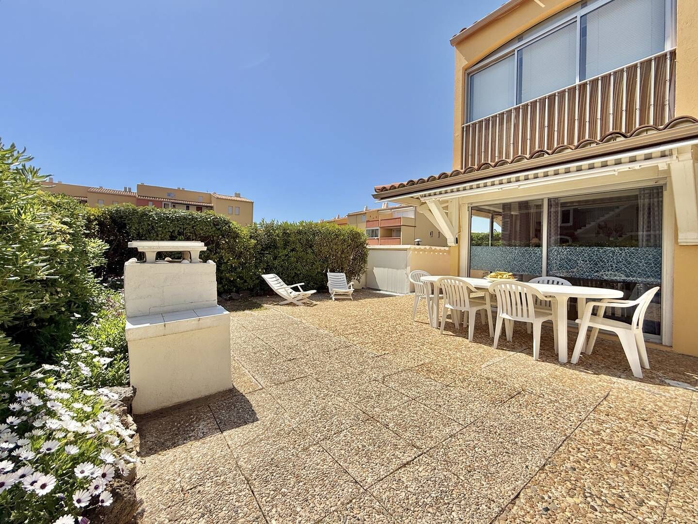 Eine Terrasse mit weißen Stühlen und einem Tisch, umgeben von einem gefliesten Boden und einem Heckenrand.