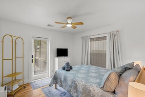 Une chambre à coucher comprenant un lit, un téléviseur, un ventilateur de plafond, une fenêtre avec des stores et une porte.