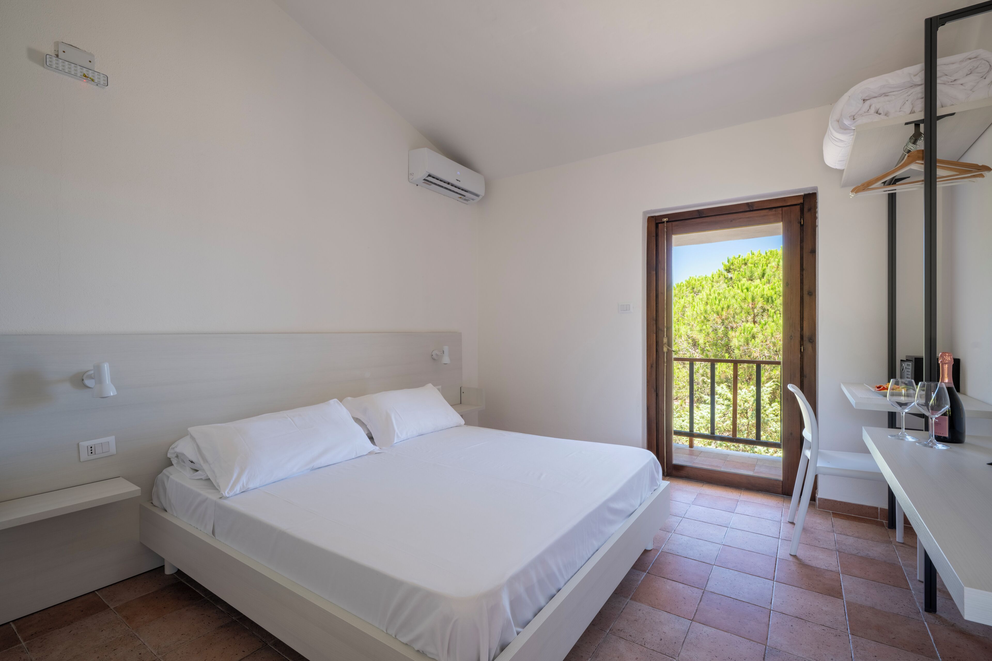 Un dormitorio con cama, escritorio y balcón con vistas a vegetación.