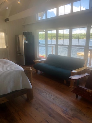 Ein Zimmer mit einem Bett, einer Couch, Holzboden, einem Kühlschrank und Seeblick durch große Fenster.