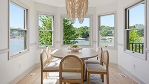 A dining room with a round table, four chairs, a chandelier, and large windows with a view of a lake and trees. - Casa del Sol | Pickle ball court| Fire pit | Dock | Sleeps 10 (Mooresville)