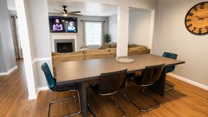 Een eetgedeelte met een lange tafel, vier stoelen, een klok aan de muur en een televisie op de achtergrond.