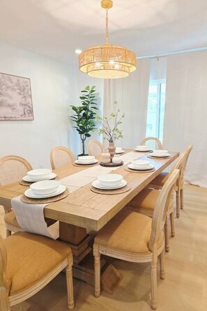 A dining room with a wooden table set for six, surrounded by upholstered chairs, a hanging light fixture, and a potted plant.