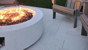 En rund eldstad med en brasa, omgiven av två Adirondack-stolar, och en ljusslinga i bakgrunden.