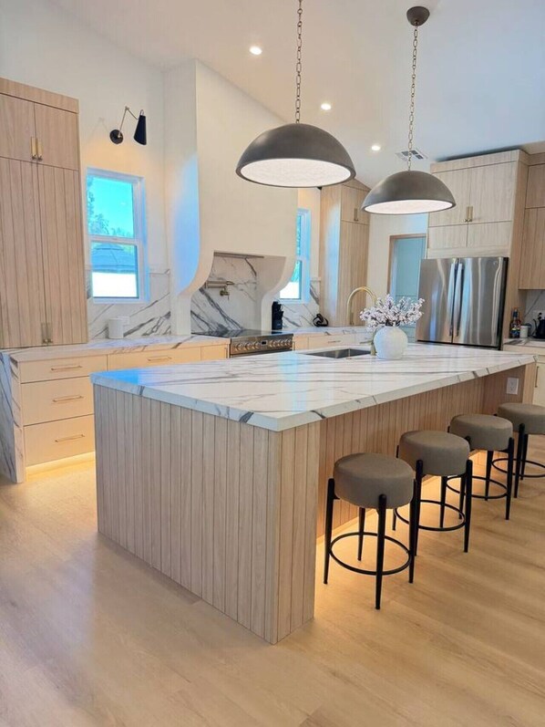 Eine moderne Küche mit Kochinsel, Barhocker, Pendelleuchten und einem Edelstahl-Kühlschrank.
