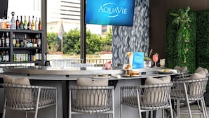 Zona de bar con una televisión mostrando 'AQUAVIE', taburetes de mimbre, barra con diferentes botellas y una pared verde con plantas.