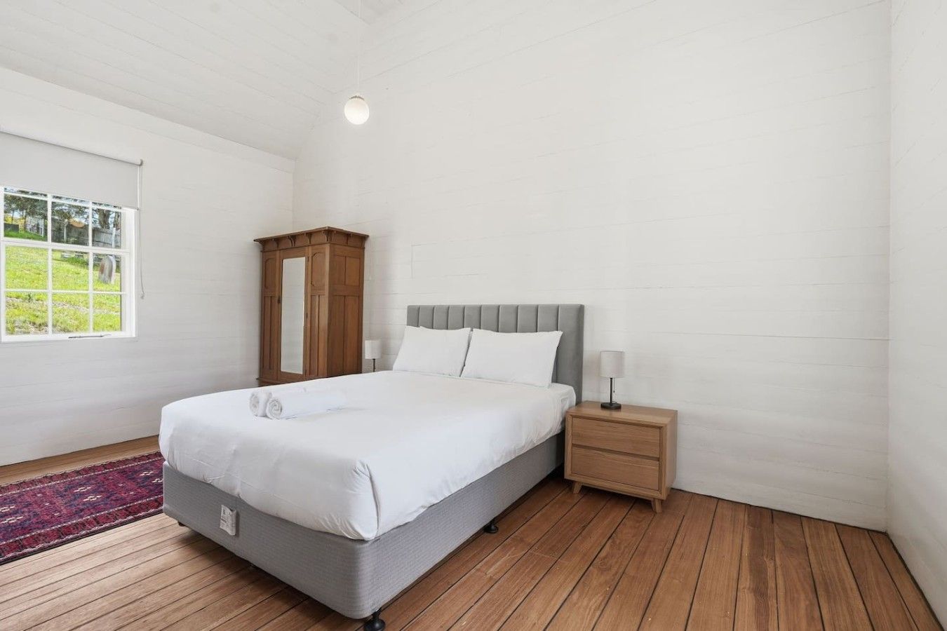 A bedroom with a bed, bedside table, nightstand, wardrobe, and a window with a view of greenery.
