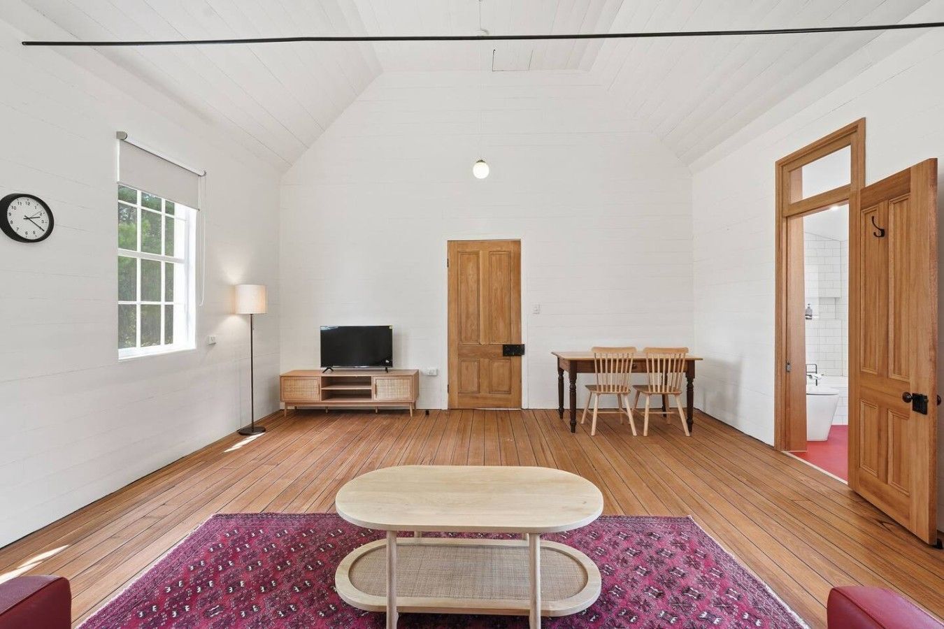 Una habitación con una televisión sobre un soporte de madera, una mesa de comedor con sillas, una puerta de madera y un reloj en la pared.