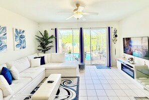 A modern living room with a sofa, TV, ceiling fan, and a view of a pool. - Spacious Townhome at a Resort Near Disney with Private Pool (Kissimmee)