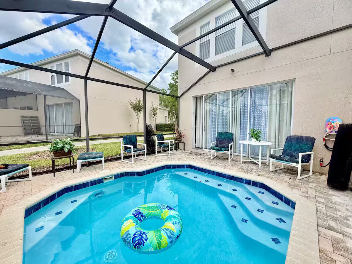 Una piscina con un flotador inflable azul y verde, rodeada de sillas de descanso blancas y un porche cubierto.