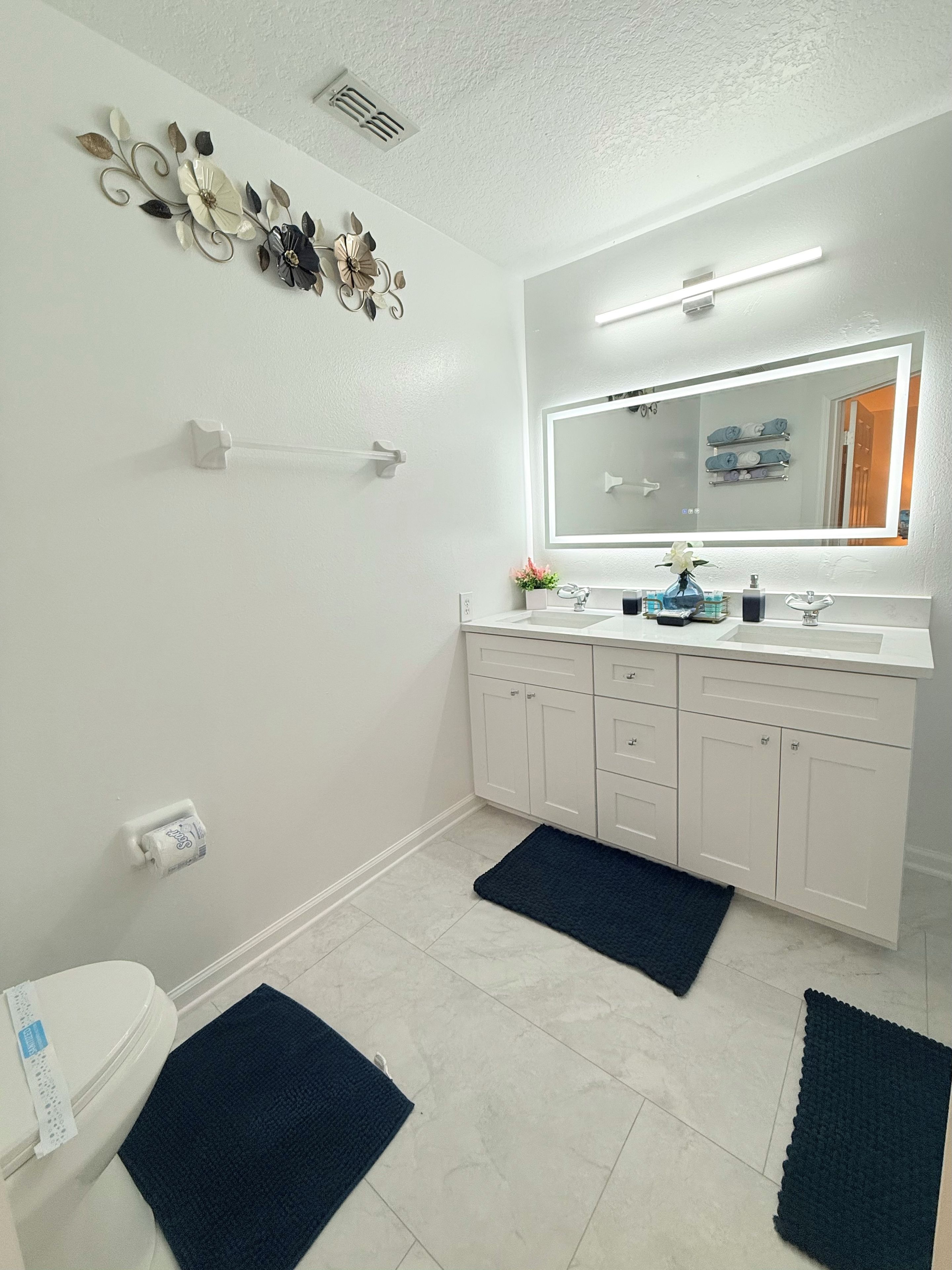 Une salle de bain avec un lavabo blanc, un miroir et des meubles de rangement, des toilettes et des œuvres d’art décoratives au mur.