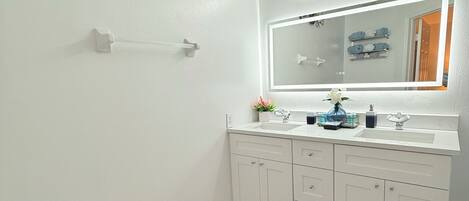 Une salle de bain avec un lavabo blanc, un miroir et des meubles de rangement, des toilettes et des œuvres d’art décoratives au mur.