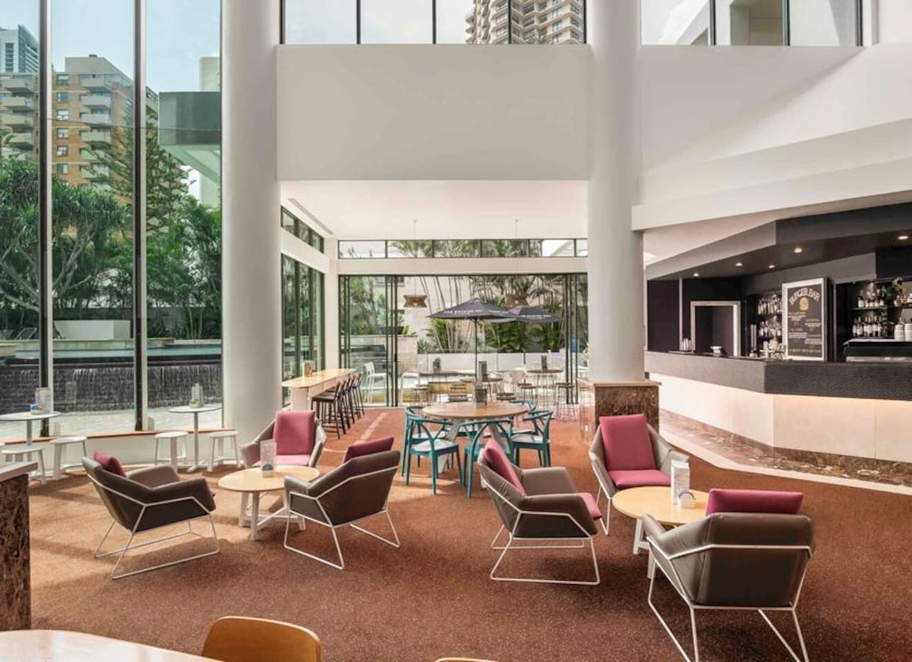 A modern hotel lobby with a high ceiling, large windows, a bar area, and seating arrangements.