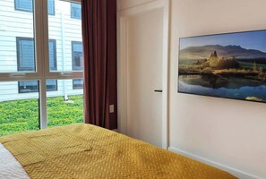 Ein Hotelzimmer mit Bett, einem fensterbelaubten Fenster und einem an der Wand montierten Fernseher, der eine Landschaft zeigt.