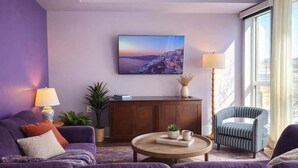 Ein Wohnzimmer mit einem violetten Sofa, einem runden Couchtisch, einem Bücherregal, einer Lampe und einem großen Fenster.