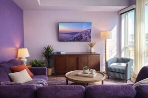 A living room with a purple sofa, a round coffee table, a bookshelf, a lamp, and a large window. - Fifa Stay 5 Min Walk Met Sleeps 5 3 Beds Gym (Philadelphia)