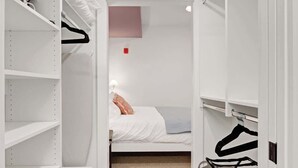 Ein begehbarer Kleiderschrank mit offenen Regalen, ein Bett mit weißer Decke und eine Kleiderstange mit Kleidung.