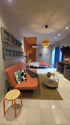 Ein modernes Wohnzimmer mit einer Couch, einem Glastisch, einem Bett mit gestreifter Bettdecke und einem Essbereich mit Hocker.