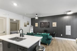 Ett modernt kök med en vit bänkskiva och en diskho i rostfritt stål, en mörk vägg med en väggmonterad TV, en grön soffa och en takfläkt.