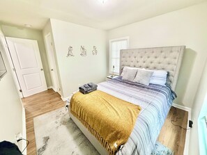 Ein Schlafzimmer mit Bett, Nachttisch, Lampe, einem Fenster mit Jalousien und einer Tür.