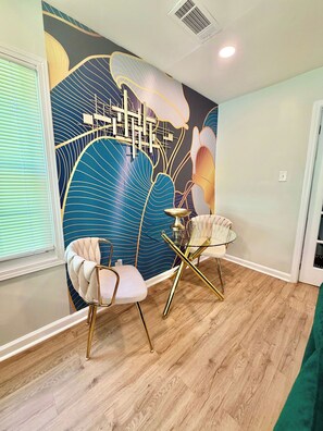 Ein Zimmer mit einem modernen Wandgemälde, zwei weißen Stühlen mit goldenen Beinen, einem Glastisch und Holzboden.