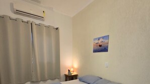 A bedroom with a bed, a nightstand, a lamp, a painting, and a window with curtains. - Flats Brisapart (Ubatuba)