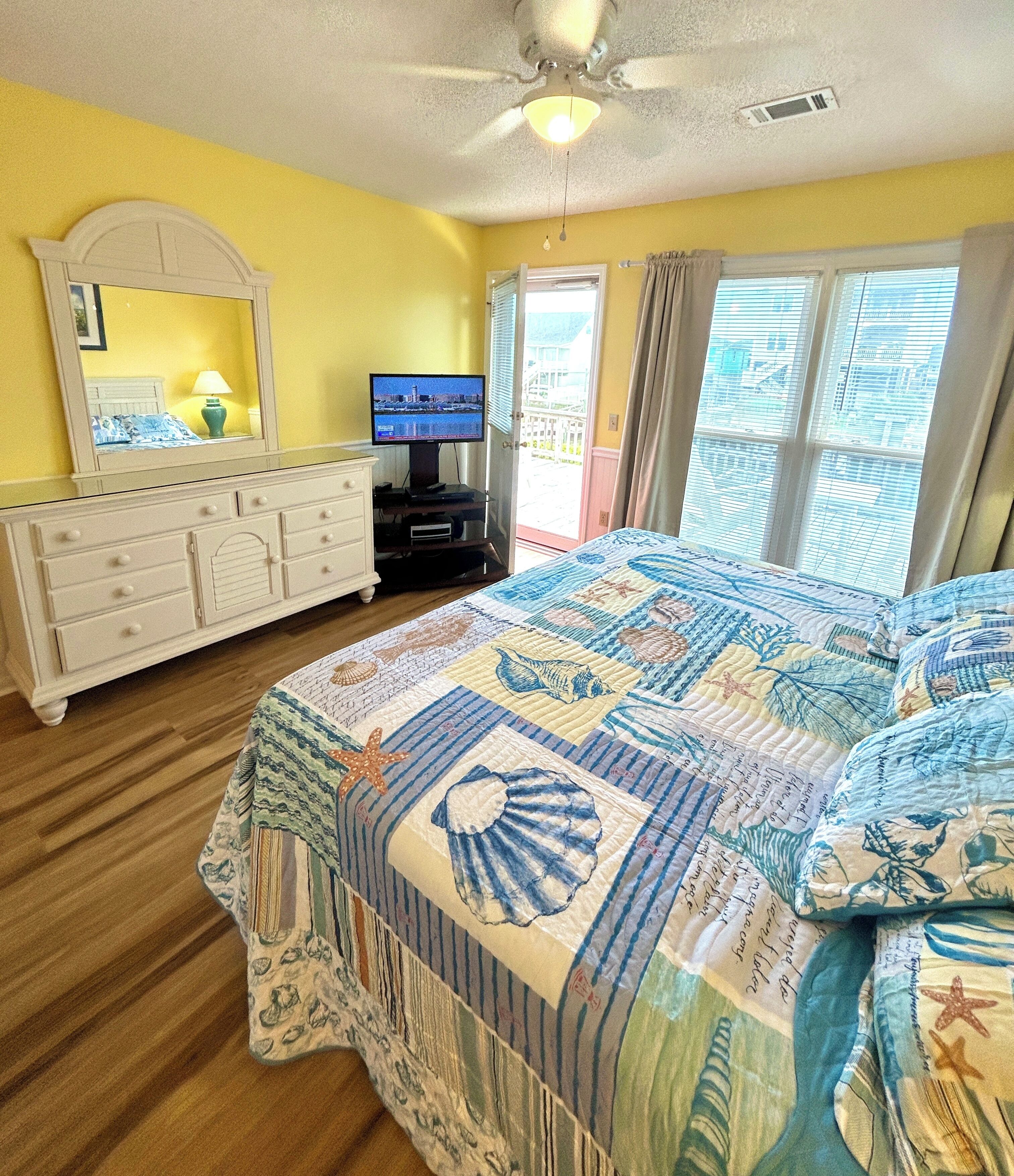 A bedroom with a bed, dresser, television, and a view of the outside.