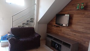 A living room with a covered sofa, a staircase, a TV mounted on a wooden wall, and a small shelf with speakers. - Quiet corner (Igarapé)