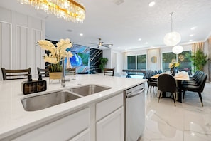 A modern kitchen with a built-in dishwasher, a large island with a sink, and a dining area with a long table and chairs. - Classy 12BR, 11.5Bath, Sleeps 32, Theater, Game Rm (Kissimmee)