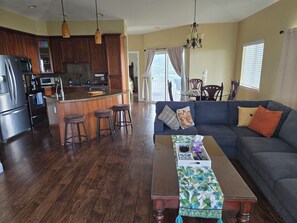 Eine moderne Küche mit einer zentralen Kochinsel, Barhocker, einem Essbereich mit Tisch und Stühlen sowie einem Wohnzimmer mit Sofa und Couchtisch.