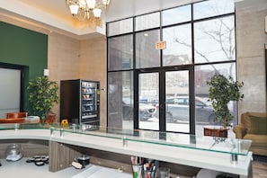 A lobby with a glass partition, vending machine, potted plants, and a view of the street outside.