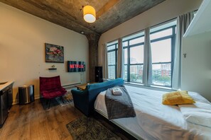 A modern hotel room with a large bed, a blue sofa, a red chair, a TV, and a view of the city through large windows.