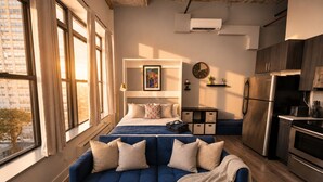 A modern bedroom with a bed, a blue sofa, a kitchenette, and a city view. - McCormick Place Modern Suite for 4, with Optional Parking (Chicago)