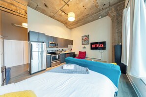 Moderni hotellihuone, jossa on pieni keittiö, sänky, taulutelevisio ja suuri ikkuna.